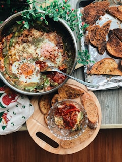 A wooden table displays a delicious breakfast spread, featuring a pan of baked eggs with herbs and possibly tomatoes. Next to it is a wooden board with toasted bread slices and a small glass bowl containing some sort of salsa or relish. Fresh green foliage decorates the scene, adding a touch of nature to the setup. A festive towel with Christmas motifs lies on the side.
