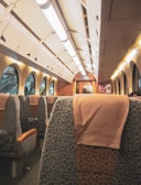An interior view of a train compartment with cushioned seats covered in patterned fabric. The overhead lighting is bright, and there are large windows on one side, allowing outside light to filter in. The seating arrangement appears comfortable, with light brown headrest covers.