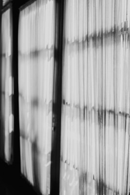 Black and white curtains hang in a row with light filtering through them, casting shadows and creating a pattern of lines and shapes on the fabric.