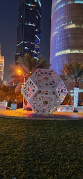 A geometric sculpture is set in a landscaped urban area with greenery and palm trees. Surrounding it are modern skyscrapers illuminated against the night sky, with one featuring intricate, illuminated patterns.