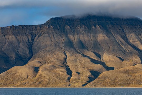 A massive rocky mountain rises sharply from the calm waters of a body of water. The mountain's surface is characterized by rugged, eroded patterns and deep vertical grooves. A thin layer of clouds partially covers the peak, adding a sense of mystery. The base of the mountain features gentle slopes and sparse vegetation.