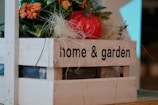 A wooden crate labeled 'home & garden' contains decorative elements such as artificial flowers and a red artificial apple. The crate is filled with green leaves and orange flowers, set against a backdrop of neutral tones.