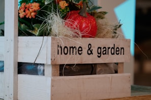 A wooden crate labeled 'home & garden' contains decorative elements such as artificial flowers and a red artificial apple. The crate is filled with green leaves and orange flowers, set against a backdrop of neutral tones.