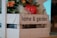 A wooden crate labeled 'home & garden' contains decorative elements such as artificial flowers and a red artificial apple. The crate is filled with green leaves and orange flowers, set against a backdrop of neutral tones.