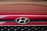 A close-up of the front grille of a red Hyundai car, displaying the Hyundai logo prominently in the center. The grille features a black and chrome mesh design.