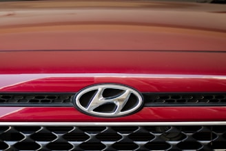 A close-up of the front grille of a red Hyundai car, displaying the Hyundai logo prominently in the center. The grille features a black and chrome mesh design.