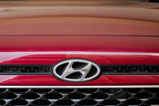 A close-up of the front grille of a red Hyundai car, displaying the Hyundai logo prominently in the center. The grille features a black and chrome mesh design.
