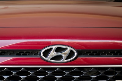 A close-up of the front grille of a red Hyundai car, displaying the Hyundai logo prominently in the center. The grille features a black and chrome mesh design.