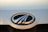 A close-up of a car emblem featuring a chrome oval shape with black and silver elements. The background appears to be a light-colored car surface with some visible specks and dirt.