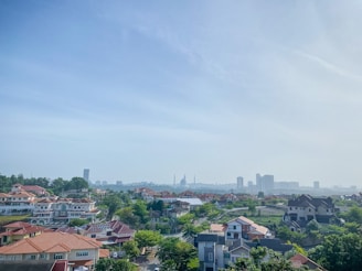 A suburban landscape features numerous houses with red-tiled roofs and lush greenery, set against a backdrop of urban skyscrapers on the horizon under a clear blue sky.