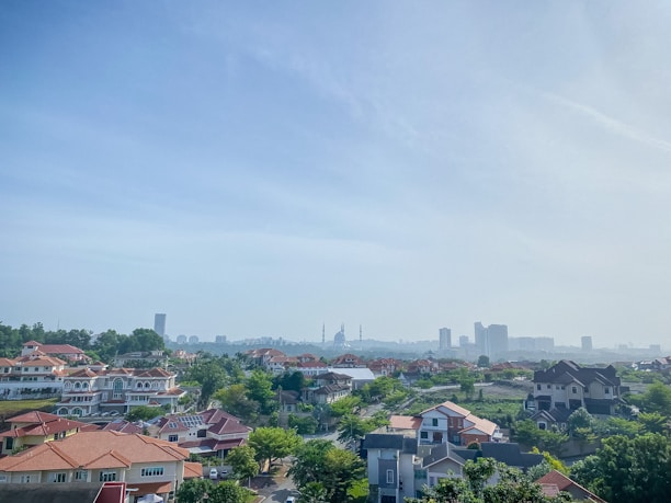 A suburban landscape features numerous houses with red-tiled roofs and lush greenery, set against a backdrop of urban skyscrapers on the horizon under a clear blue sky.