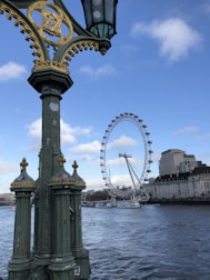 A large Ferris wheel, known as the London Eye, is prominently featured against a clear blue sky. Nearby, an ornate lamppost with gold and green accents stands on a bridge overlooking a river. The water is calm, and in the background, modern and historical buildings line the shoreline.