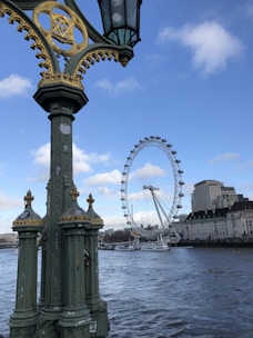 A large Ferris wheel, known as the London Eye, is prominently featured against a clear blue sky. Nearby, an ornate lamppost with gold and green accents stands on a bridge overlooking a river. The water is calm, and in the background, modern and historical buildings line the shoreline.