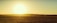 A vast open field stretches under a clear sky with a bright, low-hanging sun on the horizon, casting a warm golden hue over the landscape. An irrigation system extends diagonally across the field, consisting of metal structures with wheels that suggest modern agricultural practices.