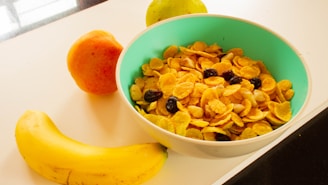 A bowl filled with cornflakes and dried fruit sits on a table. Surrounding the bowl are a banana, an orange, and a green pear.