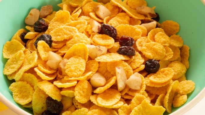 A close-up view of a bowl filled with cornflakes, mixed with nuts and raisins. The cornflakes are golden and crispy, while the nuts appear to be sliced, adding texture. The raisins are dark and scattered throughout, providing contrast.