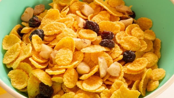 A close-up view of a bowl filled with cornflakes, mixed with nuts and raisins. The cornflakes are golden and crispy, while the nuts appear to be sliced, adding texture. The raisins are dark and scattered throughout, providing contrast.