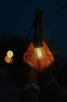 A pair of hands gently cradles a glowing light bulb, creating a warm and intimate atmosphere. The background is dark, highlighting the soft glow of the bulb and hands.