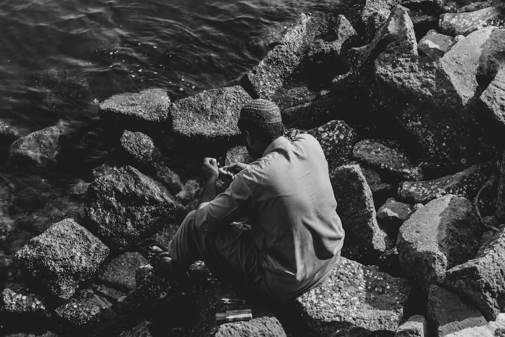 man in black jacket and pants sitting on rock near body of water