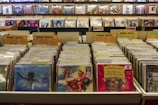 A record store display features numerous vinyl records organized in bins with labels like 'Indie Rock' and 'Punk/Hardcore.' Album covers are visible on the front of some records, while others are partially obscured. The background has shelves with more records displayed upright, and a sign that reads 'DJ Headquarters' is partially visible at the top.