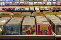A record store display features numerous vinyl records organized in bins with labels like 'Indie Rock' and 'Punk/Hardcore.' Album covers are visible on the front of some records, while others are partially obscured. The background has shelves with more records displayed upright, and a sign that reads 'DJ Headquarters' is partially visible at the top.