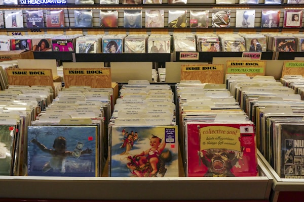 A record store display features numerous vinyl records organized in bins with labels like 'Indie Rock' and 'Punk/Hardcore.' Album covers are visible on the front of some records, while others are partially obscured. The background has shelves with more records displayed upright, and a sign that reads 'DJ Headquarters' is partially visible at the top.
