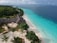 An aerial view of a tropical beachfront resort with lush greenery, thatched-roof buildings, and a clear turquoise ocean stretching alongside a white sandy beach. The resort is surrounded by well-maintained gardens with palm trees and paths leading to different parts of the area.