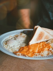 A plate of breakfast food featuring two slices of toasted bread, a serving of hash browns covered with a slice of melted cheddar cheese, and two fried eggs. The table is subtly lit, giving a warm and cozy ambiance.