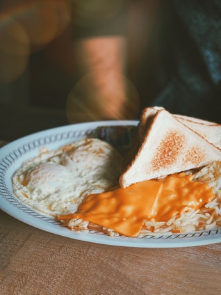 A plate of breakfast food featuring two slices of toasted bread, a serving of hash browns covered with a slice of melted cheddar cheese, and two fried eggs. The table is subtly lit, giving a warm and cozy ambiance.