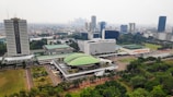 An aerial view of a large urban area featuring a mix of modern high-rise buildings and lower structures with distinctive green roofs, set against a backdrop of a sprawling cityscape. The lush greenery and landscaped gardens are visible in the foreground, contrasting with the dense urban environment.