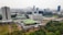 An aerial view of a large urban area featuring a mix of modern high-rise buildings and lower structures with distinctive green roofs, set against a backdrop of a sprawling cityscape. The lush greenery and landscaped gardens are visible in the foreground, contrasting with the dense urban environment.