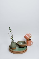 A collection of pottery items displayed against a plain background. The arrangement includes a green ceramic vase with a small plant, a green bowl placed inside a larger earthy-toned bowl, and two pink mugs stacked on top of each other. The items sit on a layered set of circular plates.