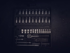 A dark toolset containing an array of metallic socket wrenches and attachments neatly arranged in a hard case. The tools have a reflective surface, and the case has defined slots for each tool, creating an orderly appearance.