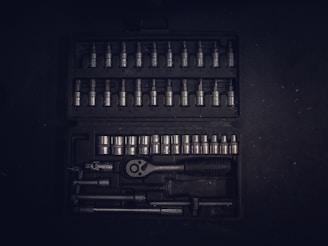A dark toolset containing an array of metallic socket wrenches and attachments neatly arranged in a hard case. The tools have a reflective surface, and the case has defined slots for each tool, creating an orderly appearance.