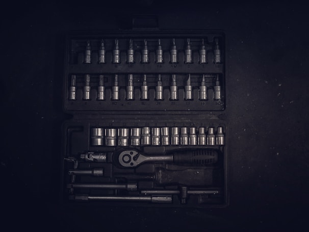 A dark toolset containing an array of metallic socket wrenches and attachments neatly arranged in a hard case. The tools have a reflective surface, and the case has defined slots for each tool, creating an orderly appearance.