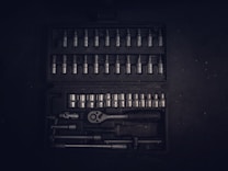 A dark toolset containing an array of metallic socket wrenches and attachments neatly arranged in a hard case. The tools have a reflective surface, and the case has defined slots for each tool, creating an orderly appearance.