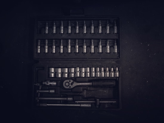 A dark toolset containing an array of metallic socket wrenches and attachments neatly arranged in a hard case. The tools have a reflective surface, and the case has defined slots for each tool, creating an orderly appearance.