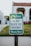 A green and white sign with the words 'Please Do Not Block The Driveway Thank You!' stands on a metal pole. The sign is set against the backdrop of a residential area, with a blurred image of a house and lawn in the background.