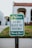 A green and white sign with the words 'Please Do Not Block The Driveway Thank You!' stands on a metal pole. The sign is set against the backdrop of a residential area, with a blurred image of a house and lawn in the background.