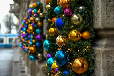 Colorful Christmas decorations with shiny baubles including gold, blue, and purple hues adorn a wall. Greenery surrounds the ornaments, and small lights add a festive sparkle. A blurred background shows a street with trees and a tram.