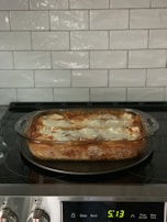 A freshly baked lasagna sits in a glass casserole dish on an electric stovetop. The kitchen backsplash features white subway tiles.