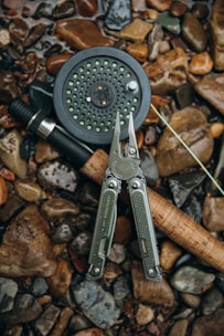 A fishing reel with green details lies on a bed of wet, multicolored pebbles. Next to it is a multi-tool with pliers opened, featuring a textured grip and metallic finish. The fishing line is partially visible.