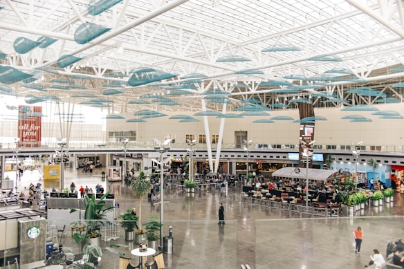 A spacious and modern airport terminal with large glass windows allowing natural light to fill the area. The ceiling features artistic teal accents hanging from white structural beams. The terminal includes shops and cafes with several people sitting or walking through, some with luggage. Potted plants are placed throughout the space, adding a touch of greenery.