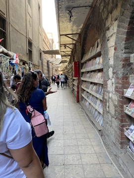 A narrow alleyway lined with books on shelves along the brick wall to the right. People are walking and browsing, with some carrying backpacks and bottles. The setting has an urban, rustic feel with a mixture of stone and concrete structures.