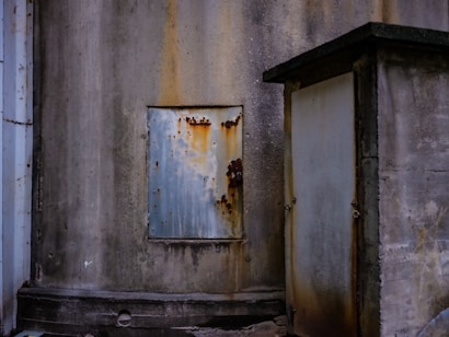 Weathered and rust-stained surfaces of an industrial structure with a metallic panel and a utility box on a concrete wall. The presence of rust and discoloration suggests age and exposure to the elements.