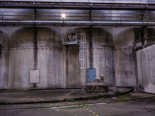 An industrial setting with large, weathered concrete walls showcasing a metal ladder leading to a platform. The area is dimly lit, with an overhead light illuminating part of the scene. A small door and various geometric structures add to the utilitarian atmosphere.