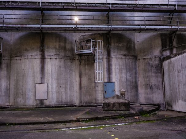 An industrial setting with large, weathered concrete walls showcasing a metal ladder leading to a platform. The area is dimly lit, with an overhead light illuminating part of the scene. A small door and various geometric structures add to the utilitarian atmosphere.