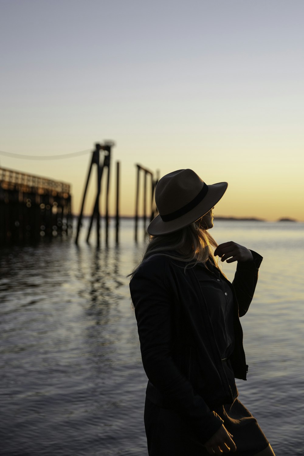 日没時にビーチに立つ黒いジャケットと茶色の日よけ帽をかぶった女性