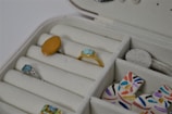 A jewelry box with multiple compartments contains rings and other accessories. There are rings with different colored stones, including a blue and an orange one. Nearby, multicolored, abstract-patterned discs are also visible.