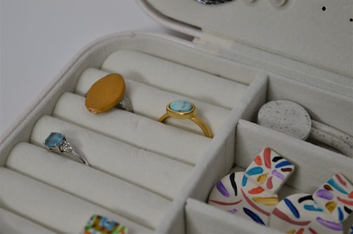 A jewelry box with multiple compartments contains rings and other accessories. There are rings with different colored stones, including a blue and an orange one. Nearby, multicolored, abstract-patterned discs are also visible.